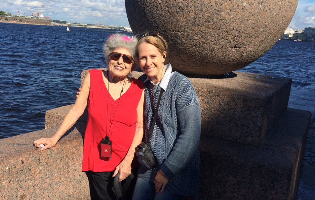 On a sunny day in Russia, Susannah and her mom, Barbara, lean against a three-foot-high granite wall beside the Volga River — a carved ball of granite balances on the corner behind them. Barbara wears a loose, bright red vee-necked sleeveless blouse, and a hearing device hangs from a cord around her neck. Her gray hair is swept away from her face and is held with bright pink flowers. She is beaming at the camera and rocks, as she used to call them, “black lips” — dark lipstick. Barbara has on dark sunglasses. Susannah wears a blue textured sweater with a camera slung across her chest. She has her arm around her mom and smiles brightly at the camera.