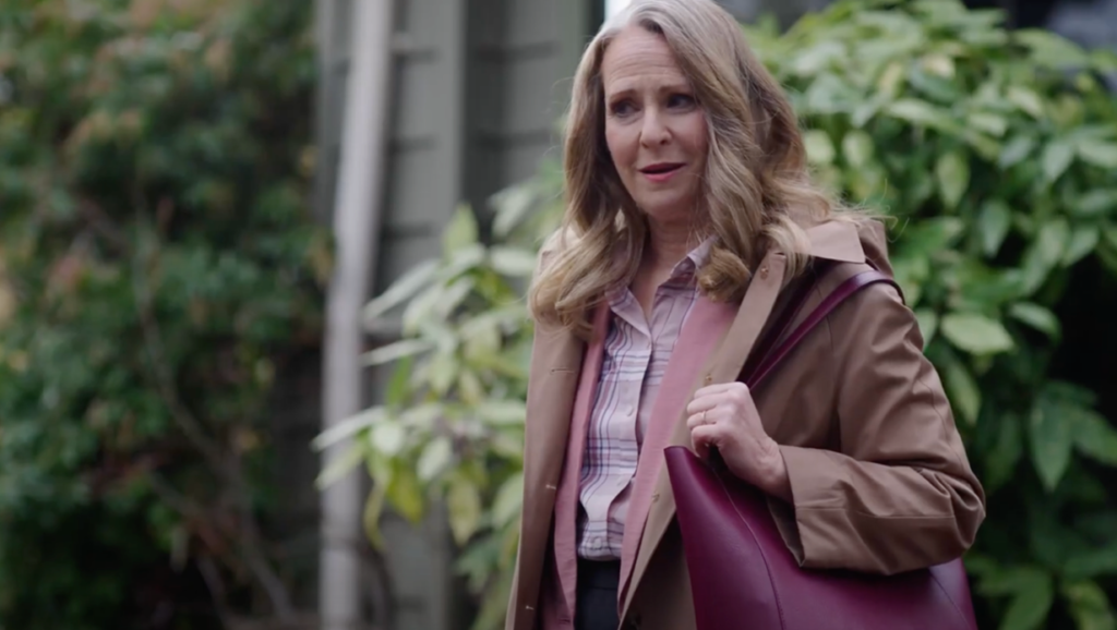 A woman in a lilac trench coat stands in from of a green leafy background. She has a pensive look and a burgundy bag slung over her shoulder.