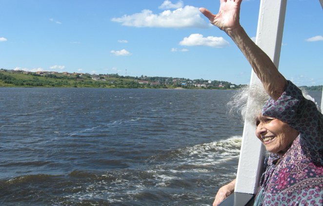On the balcony of a ship, brightly smiling and squinting in the sun, an elderly woman stretches out her left hand in greeting. She wears a multicolored, patterned blouse, and wisps of her kinky gray hair blows in the breeze. Nearby the coastline is green with groupings of red roofed buildings here and there. The sky is blue with sporadic billowy clouds.