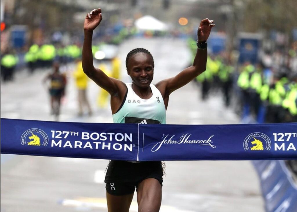 A foot-wide, navy blue finish line tape with text reading “One hundred twenty-seventh Boston Marathon” beside a yellow unicorn, is about to be broken by a dark skinned woman in a light green tank top with the letters O A C on the front. Her arms are raised.
