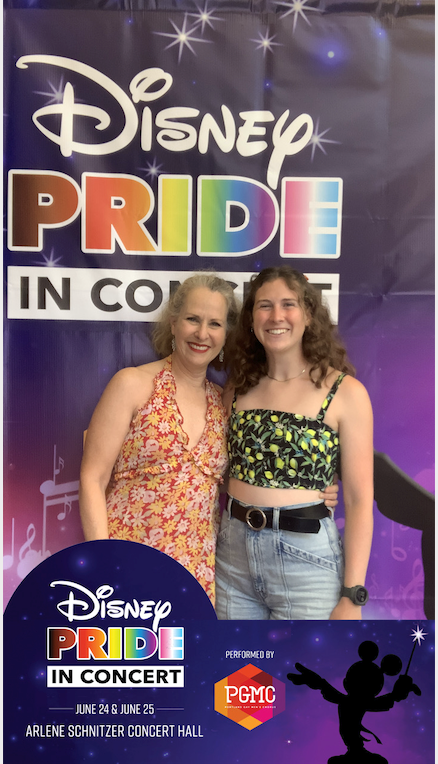 In front of a live concert step-and-repeat, Susannah, smiles in a flowered haltered dress and has her arm around her daughter's waist, a younger woman in a cropped, cropped, floral print top. Behind them, a sign reads Disney Pride In Concert. “Pride” is written in block letters filled with rainbow colors.