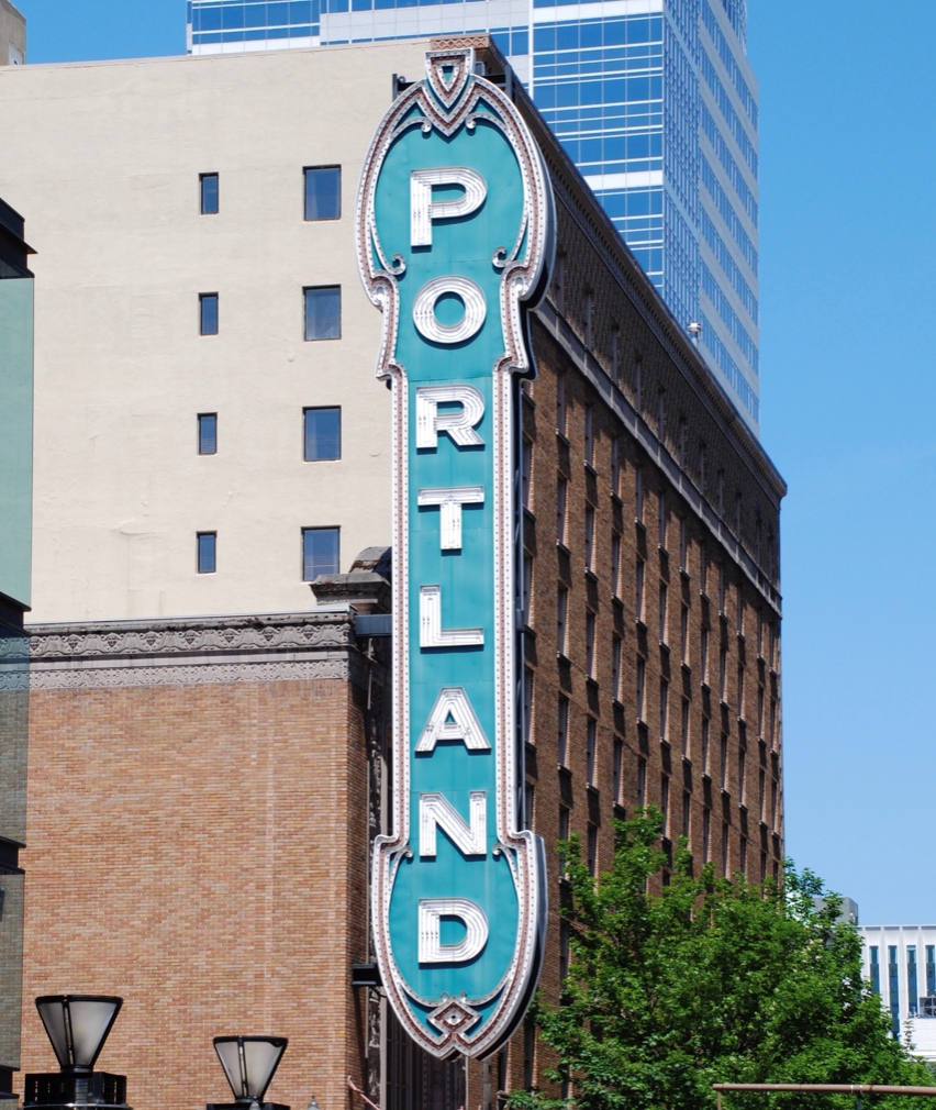 Daytime, a vertical sign on a green background with old-timey marquee lighting all around reads: PORTLAND in white block letters that light brightly at night.