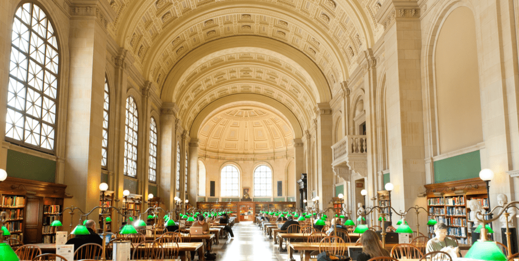 Alt ID: Bates Hall at the Boston Public Library, built 1888-1895. At left, light streams from massive arch-topped paned windows into a grand rectangular study hall with a barrel-vaulted white Ohio sandstone ceiling at the end. The room is two hundred and eighteen feet long and resembles a Roman basilica. The room is divided by two rows of rectangular wooden study tables on either side of the long walls, creating an aisle down the center. Eight curved back wooden chairs sit at each table, some occupied. A brass green glass banker’s style lamp sits atop each table.