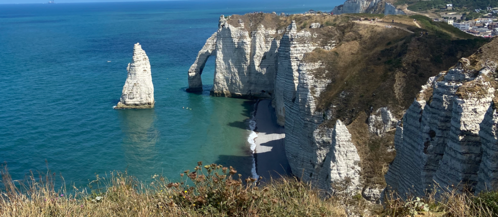 The Alabaster Coast on the Normandy Coast in France; A variegated ocean of blues and greens laps onto a narrow beach, behind which stand white, horizontally striated cliffs. One of the cliffs has eroded in a way that’s created a natural tunnel. A lone tower of white emerges from the sea in front of that tunnel.