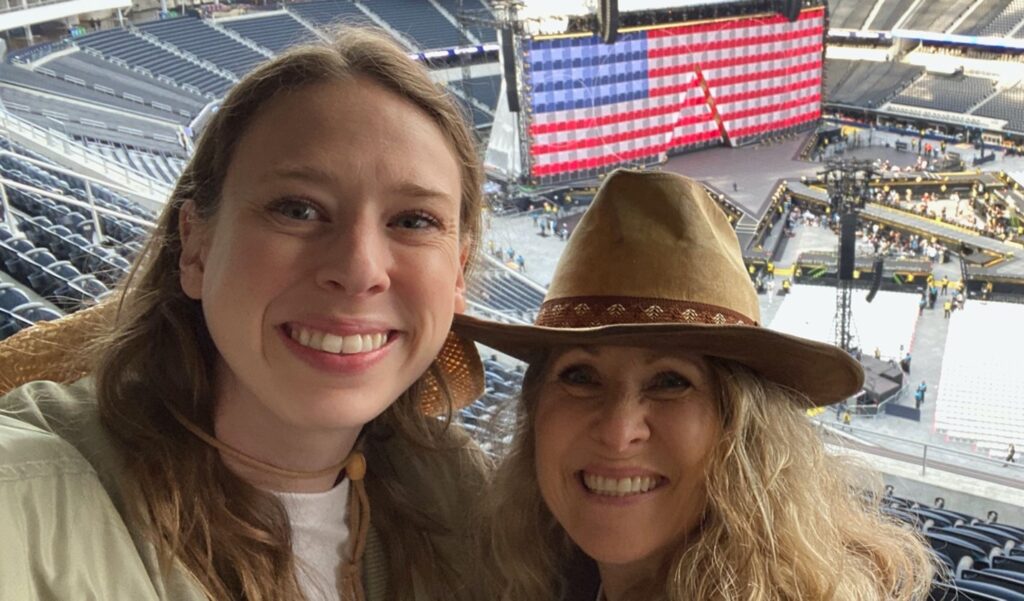 A selfie of Susannah and her daughter smiling broadly in a massive football stadium that is slowly filling with fans, about three spectator sections above the field. Susannah wears a cowboy hat and an American flag provides the backdrop for Beyoncés stage.
