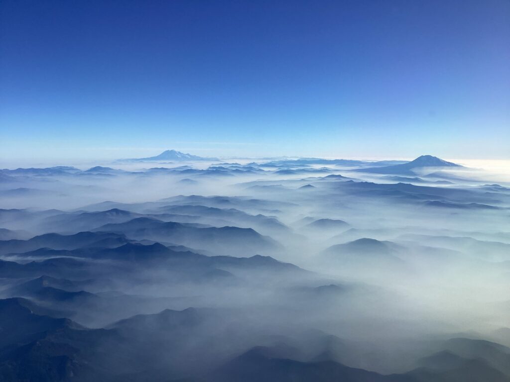 A sweeping mountain landscape viewed from above, with layers of dark blue ridgelines emerging through soft white mist. Two distant volcanic peaks rise above the clouds beneath a clear, vivid blue sky.