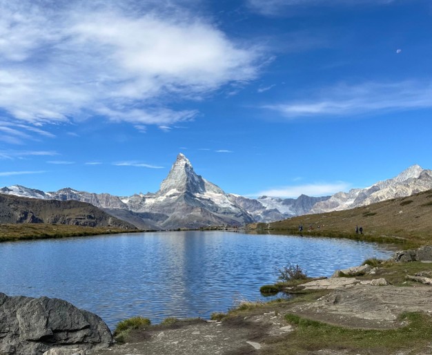 View across a clear alpine lake toward the Matterhorn, its distinctive peak rising above rugged mountains beneath a bright blue sky with wispy clouds.